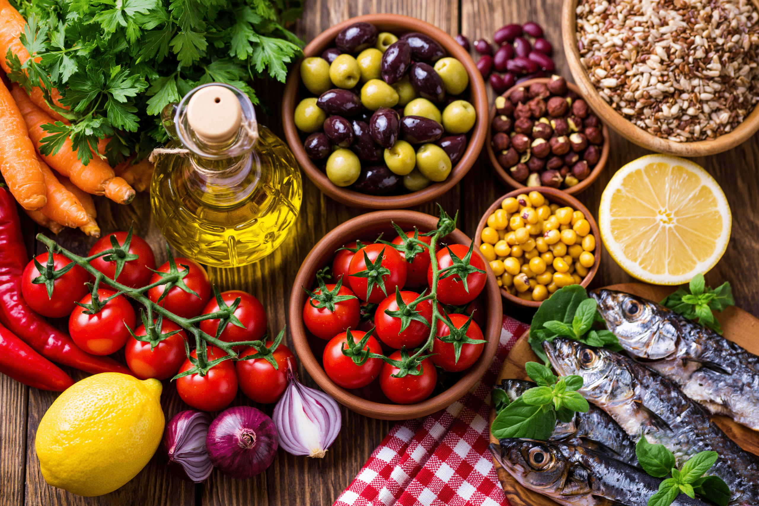 A vibrant flat lay of Mediterranean diet foods including fresh vegetables, fruits, whole grains, olives, olive oil, fish, and legumes on a rustic wooden table.